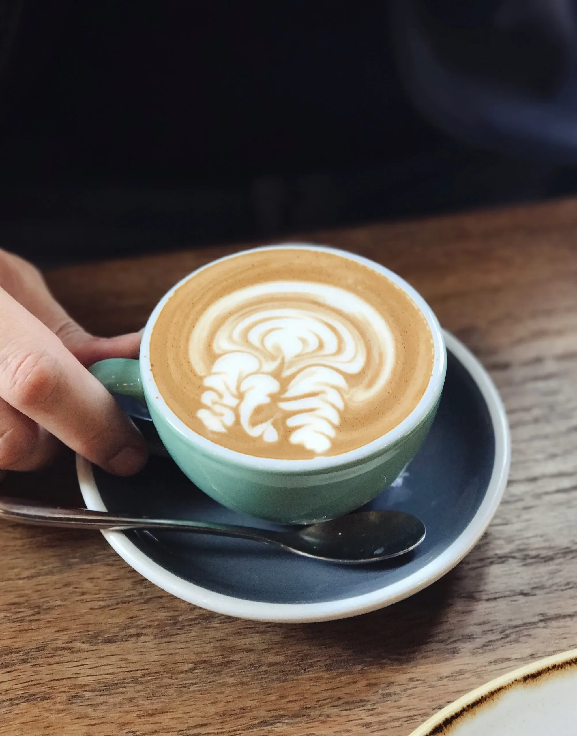 A flat white with leaf latte art in a sage-green cup on a wooden café table