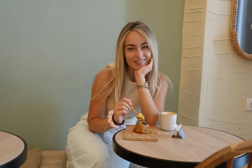 A guest at a round wooden table at Cookie Beacon Kralja Petra in Belgrade with a cookie on a board and a cappuccino, beside the sage-green wall and a raffia mirror