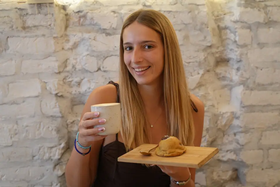 A guest holding a speckled ceramic cup and a wooden board with a chunky cookie on it, against the whitewashed brick wall at Cookie Beacon Kralja Petra in Belgrade