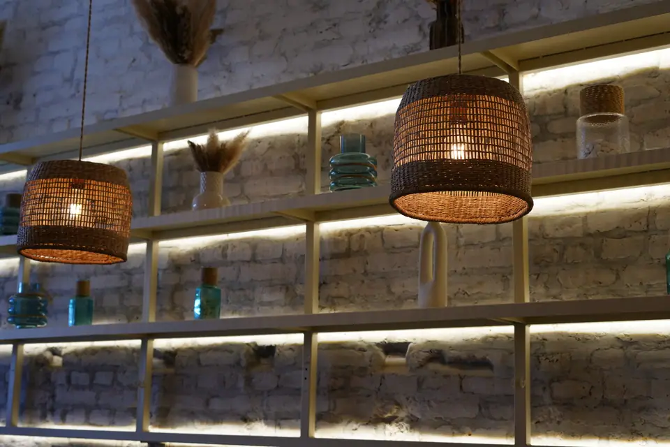 Backlit shelves of glass bottles and dried grasses against a whitewashed brick wall at Cookie Beacon Kralja Petra in Belgrade, with woven rattan pendant lamps hanging in front