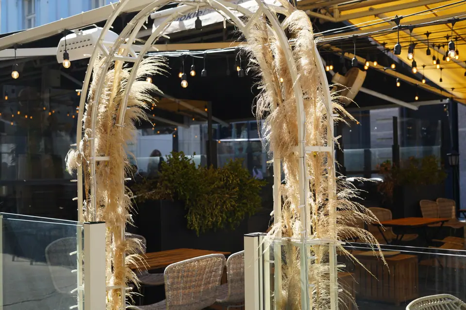 A white arch dressed with pampas grass framing the entrance to the terrace at Cookie Beacon Kralja Petra in Belgrade, with festoon lights strung under the awning behind