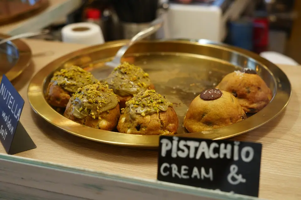 A brass tray of chunky cookies topped with pistachio cream and chopped pistachios at the counter of Cookie Beacon Kralja Petra in Belgrade, beside chalk labels reading Pistachio Cream and Nutella Maldon Salt