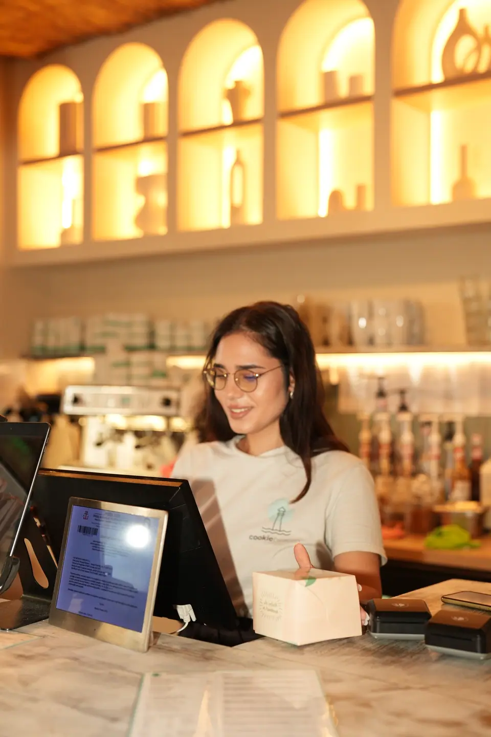 A Cookie Beacon staff member holding a takeaway box at the marble counter of the Amzei café in Bucharest, beside the card terminals and under a row of lit arched alcoves holding ceramic vases