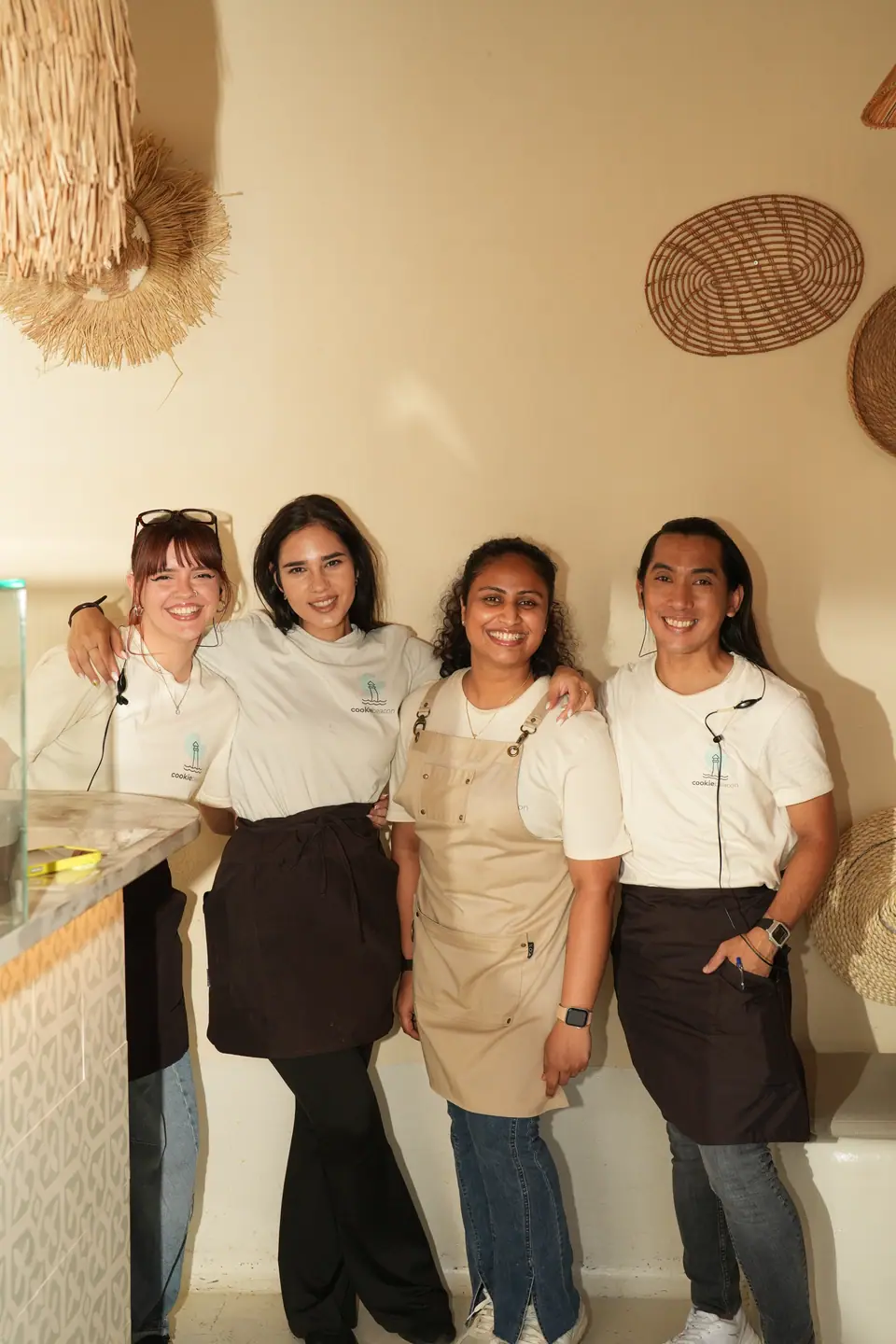Four of the Cookie Beacon team in cream t-shirts and aprons at the end of the counter in the Amzei café in Bucharest, in front of a wall of woven raffia discs