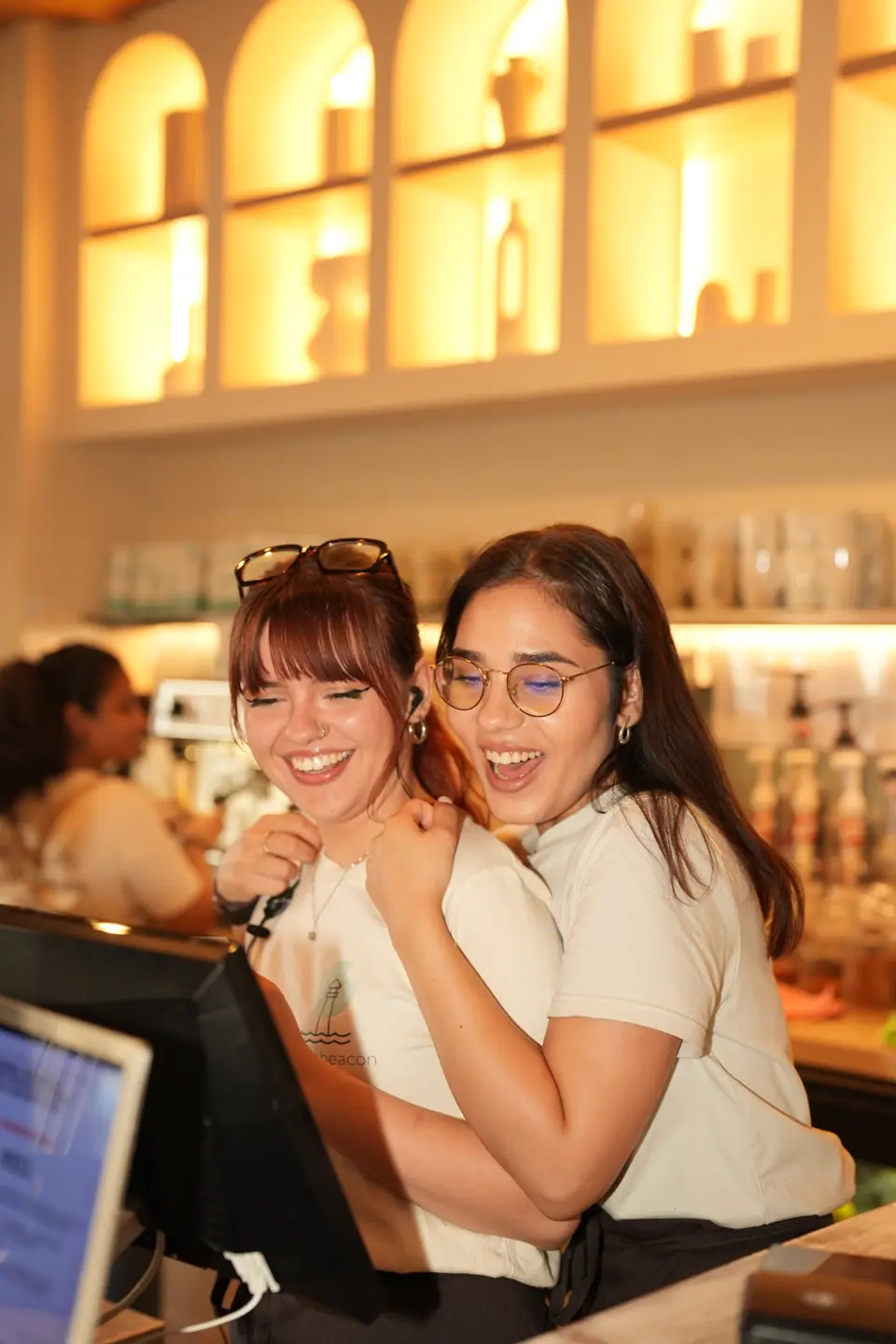 Two Cookie Beacon staff in branded t-shirts laughing behind the till at the Amzei café in Bucharest, with the lit arched alcoves above them