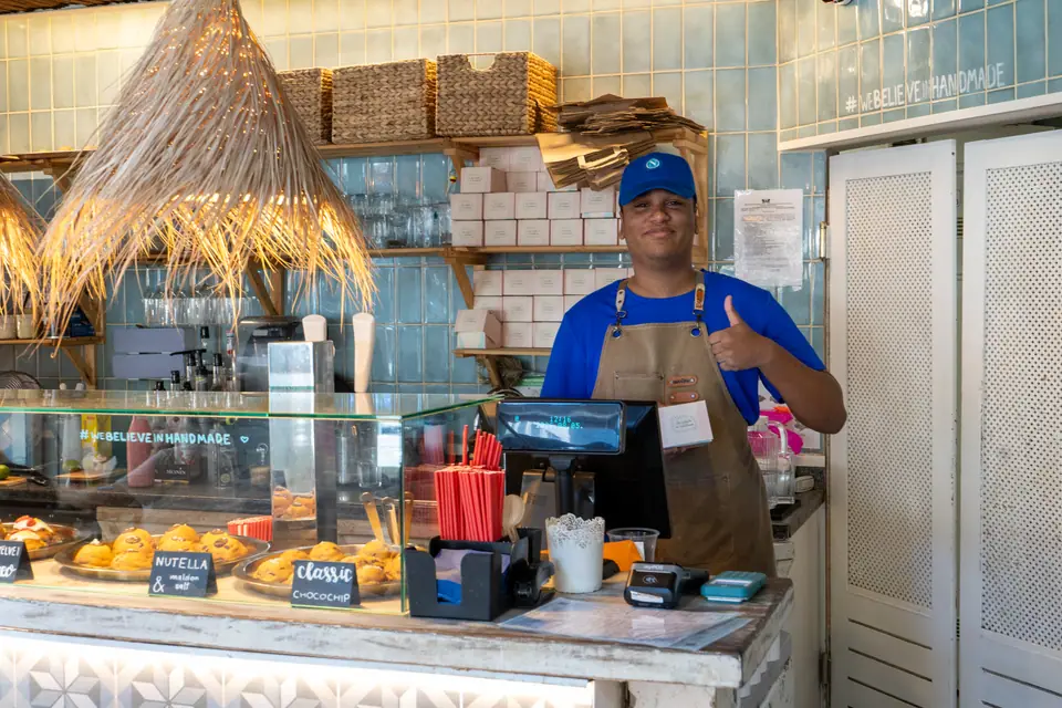 A Cookie Beacon barista in a blue cap and apron giving a thumbs up behind the counter in Budapest