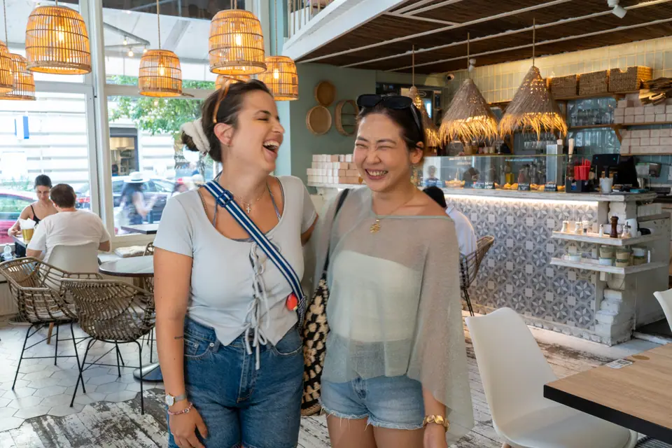Two friends laughing together inside Cookie Beacon Budapest, the counter and rattan lamps behind them