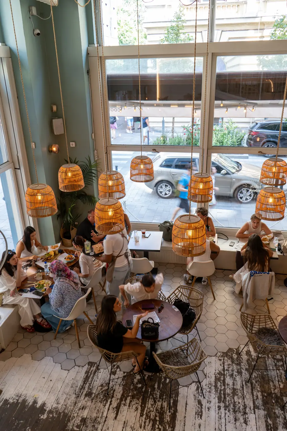 Guests at brunch beside the tall street windows of Cookie Beacon Budapest, seen from the mezzanine above
