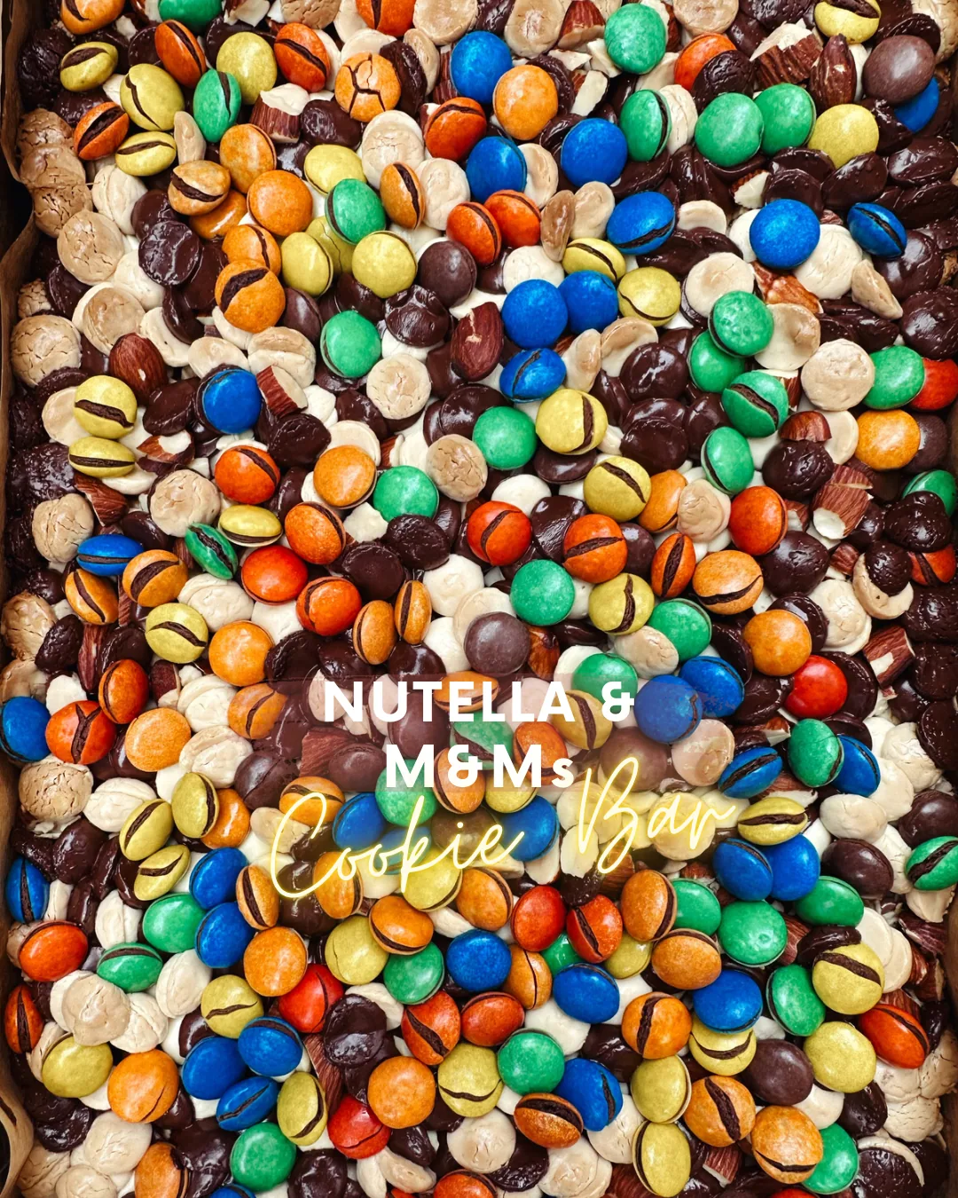 A tray of Nutella cookie bar covered edge to edge with M&M's, dark and white chocolate chips and chopped almonds
