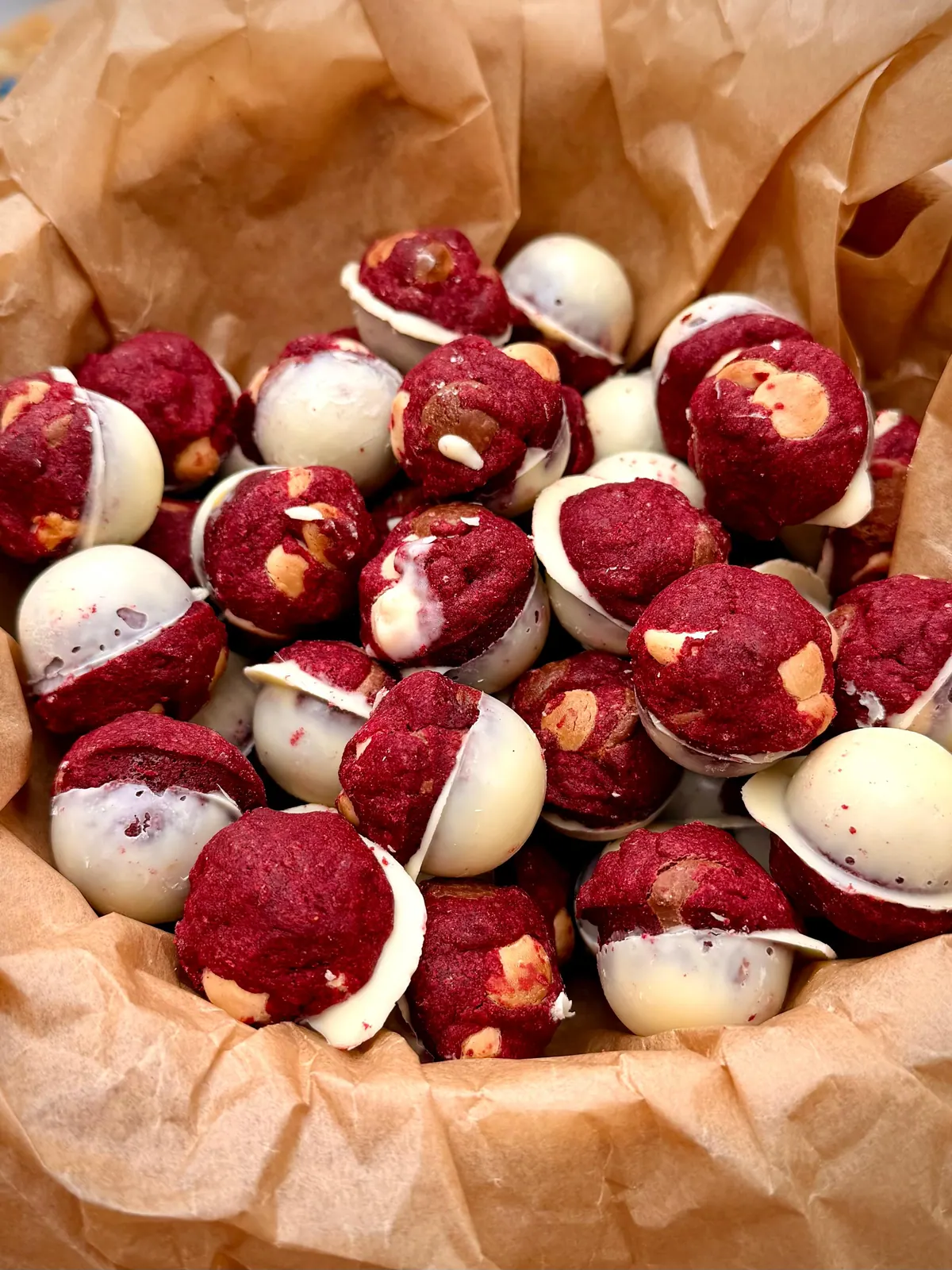 Red velvet cookie balls studded with white and milk chocolate chips, each one half-cased in a white chocolate shell, piled in brown parchment paper
